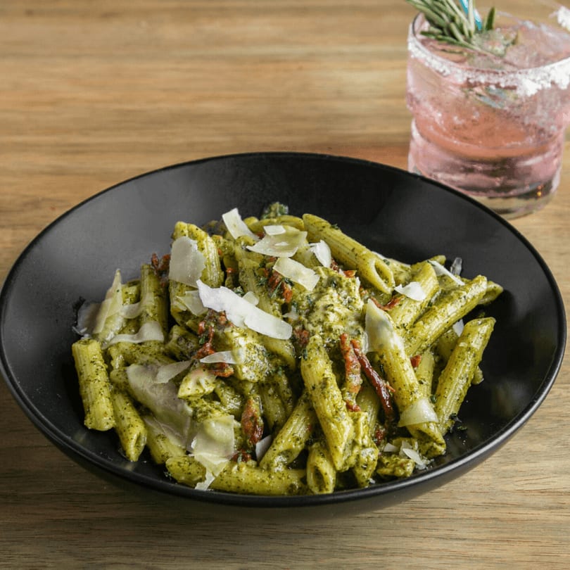 Penne pasta with pesto, sun-dried tomatoes, and parmesan cheese, with a pink cocktail.