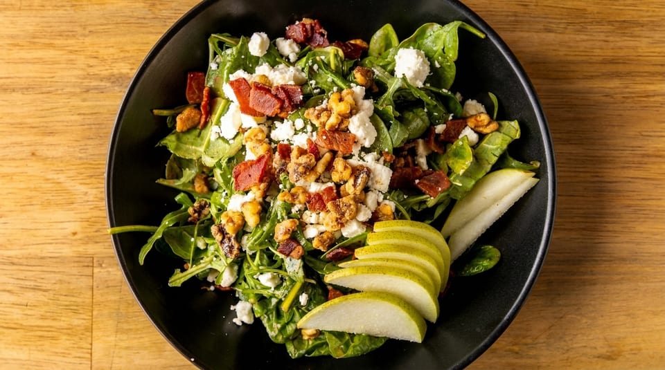 A vibrant salad with spinach, arugula, bacon, walnuts, feta cheese, and sliced pears in a black bowl.