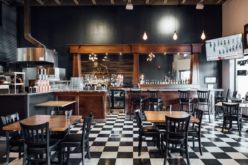 Restaurant interior with black and white checkered floor, wooden tables, black chairs, and a full bar.