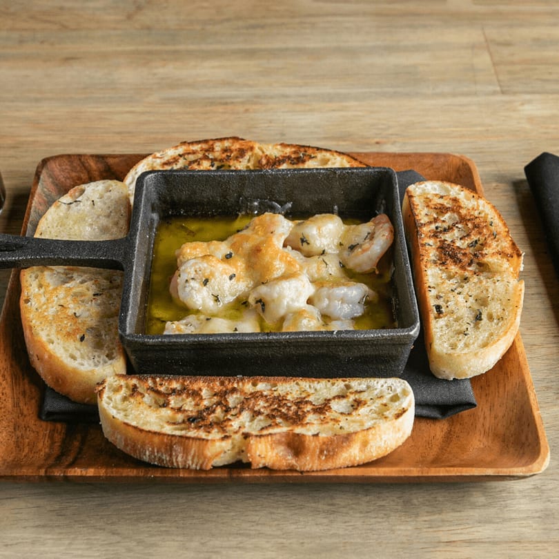 Shrimp scampi in a cast iron dish with toasted bread slices on a wooden tray.