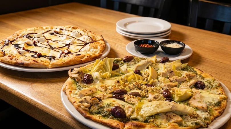 Two pizzas on a wooden table, one with pesto, chicken, olives, and artichokes, the other with cheese and balsamic glaze.
