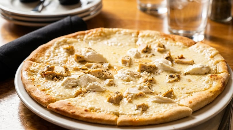 White pizza with chicken and ricotta cheese on a white plate, with a black napkin and glasses in the background.