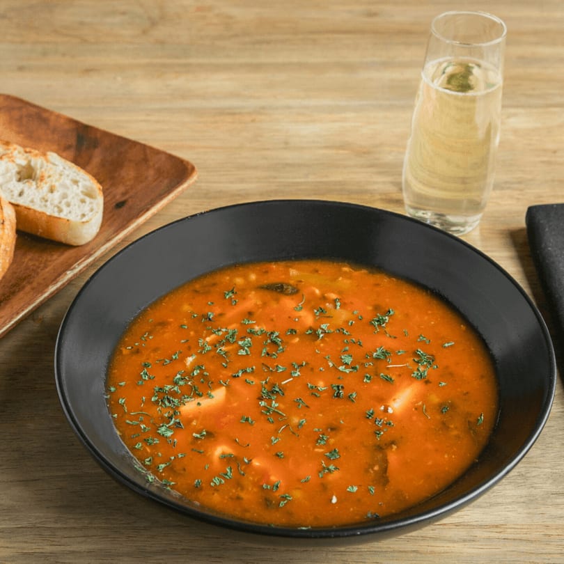 A black bowl of tomato soup with herbs, bread slices, and a glass of white wine.
