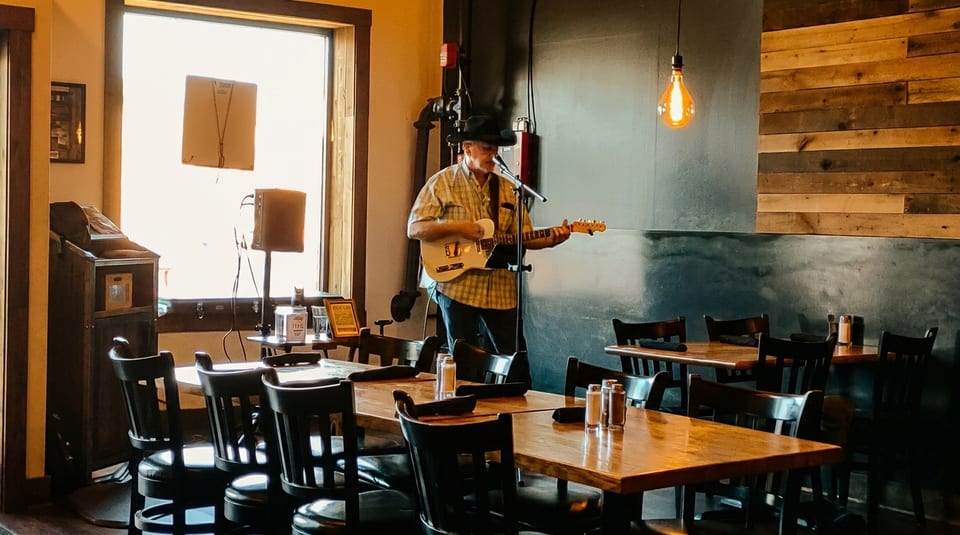 Musician in a hat playing guitar and singing in a restaurant with wooden tables and chairs.