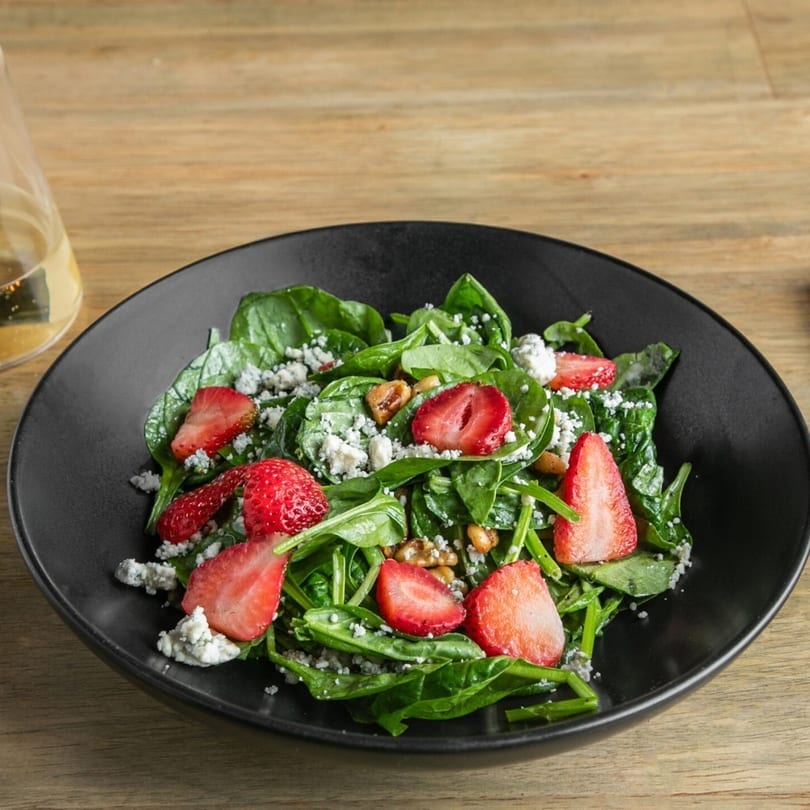 Spinach salad with sliced strawberries, crumbled blue cheese, and nuts in a black bowl.