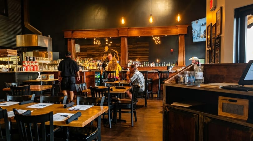 Restaurant interior with wooden tables, a bar with patrons, and a server walking through the dining area.