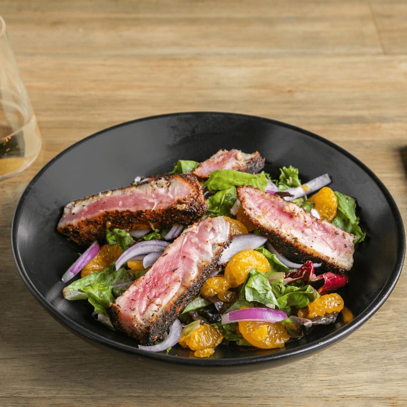 Seared tuna salad with mixed greens, red onion, and mandarin oranges in a black bowl.